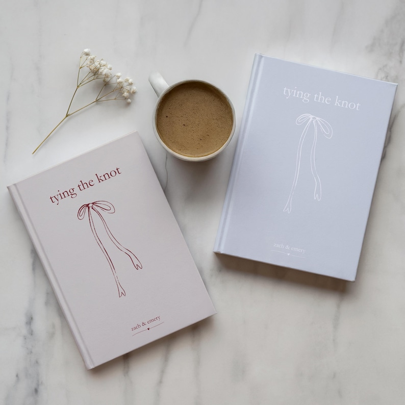 Puede incluir: Dos libros de tapa dura con el texto "tying the knot" y una ilustraci&oacute;n de lazo. Un libro es de color gris claro con texto e ilustraci&oacute;n burdeos, el otro es azul claro con texto e ilustraci&oacute;n blancos. Tambi&eacute;n hay una taza de caf&eacute; blanca y una ramita de flores blancas.