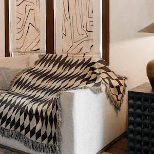 May include: A black and beige geometric patterned throw blanket with fringe detail draped over a light gray sofa. The blanket's design features a repeating diamond pattern. Decorative pillows and window treatments are visible in the background.