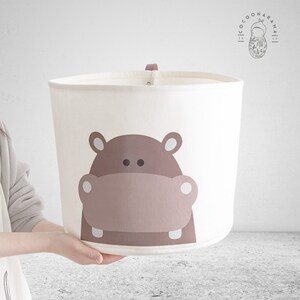 May include: A white fabric storage bin featuring a cartoon hippo face in brown tones. The bin has a brown handle and is held by a person. The cylindrical bin has a simple design. The logo "COCOONARAMA" is visible in the upper right corner.