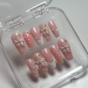 May include: A set of ten artificial nails in a clear plastic case. The nails are a light pink color with various designs, including 3D flowers, textured patterns, and gold-colored embellishments.