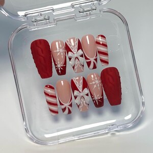 May include: A set of press-on nails in a clear plastic case. The nails feature a variety of designs, including red textured nails, white bows, snowflakes, and candy cane stripes. The nails are in a coffin shape.
