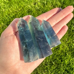 May include: Four green and blue fluorite crystal points, also known as "double terminated" crystals, are held in a hand. The crystals are pointed at both ends and have a natural, raw finish.