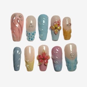 May include: A collection of ten artificial nails, each with a unique design. Colors include pink, blue, yellow, and peach, with decorative elements like swirls, dots, and flower accents. Some nails feature small, silver embellishments.