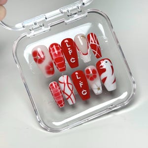 May include: A set of ten artificial nails in a clear plastic case. The nails feature a variety of designs, including red and white patterns, floral accents, and the letters "LFC" in white. Some nails have a clear base with red and white designs.