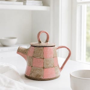 May include: A ceramic teapot featuring a hand-painted checkerboard pattern in alternating shades of dusty pink and earthy beige. The teapot has a rounded body, a curved handle, a short spout, and a lid topped with a circular loop handle.