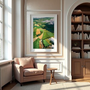 May include: A framed landscape painting depicts a patchwork of green and yellow fields, with a winding road and fluffy white clouds. The artwork hangs above a blush-colored armchair and a small wooden side table.