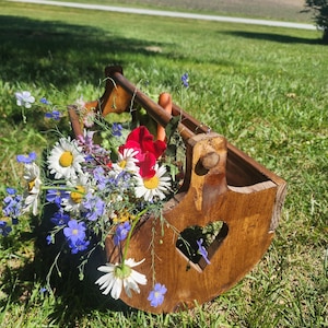 Pode incluir: Um suporte de madeira castanho com um recorte em forma de coração na lateral e uma pega superior, cheio de uma variedade de flores silvestres, incluindo margaridas brancas, pequenas flores azuis e pétalas de um vermelho brilhante.