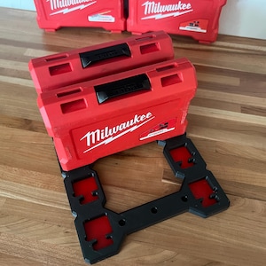 Puede incluir: Dos cajas de herramientas Milwaukee rojas apiladas en un soporte de plástico negro con cuatro ranuras. Las cajas tienen asas negras y el logotipo de Milwaukee es visible en la parte delantera.