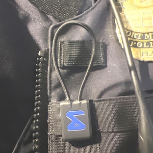 Puede incluir: Un dispositivo negro de forma cuadrada con un logotipo "S" azul unido a una correa negra. El dispositivo se lleva en un chaleco negro.