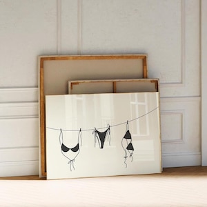 May include: A framed print of three black bikini tops hanging on a clothesline. The print is in a gold frame and is leaning against a white wall.