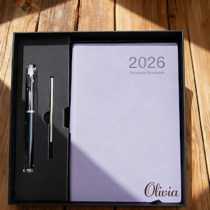 Puede incluir: Un juego de regalo en una caja negra, con un cuaderno de programación lavanda de 2026 con el nombre "Olivia" y un bolígrafo. El cuaderno está junto a un bolígrafo y un lápiz óptico de metal. El juego está sobre una superficie de madera.