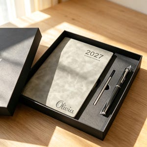Puede incluir: Un cuaderno de planificación gris 2027 con cubierta texturizada que presenta el nombre Olivia en fuente cursiva, presentado en una caja de regalo negra junto a un bolígrafo negro y un recambio de tinta metálico. La cubierta tiene un acabado mate.