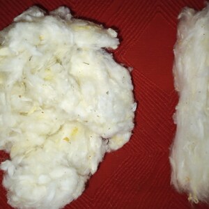 May include: Two clumps of white cotton fiber, one in a loose, rounded shape and the other in a long, thin shape, on a red textured surface.