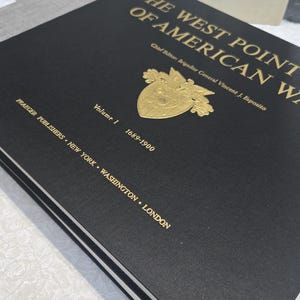 Puede incluir: Un libro de tapa dura negro titulado "THE WEST POINT OF AMERICAN WAR" con letras doradas y un emblema dorado. El volumen 1 cubre los años 1689-1900. Publicado por Praeger Publishers, con sedes en Nueva York, Washington y Londres.