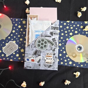 May include: A movie night gift set with a navy blue and gold star pattern, popcorn, and a CD. The set includes a pocket with newspaper print, a film reel sticker, and the text "NEVER judge a BOOK by its MOUIE".