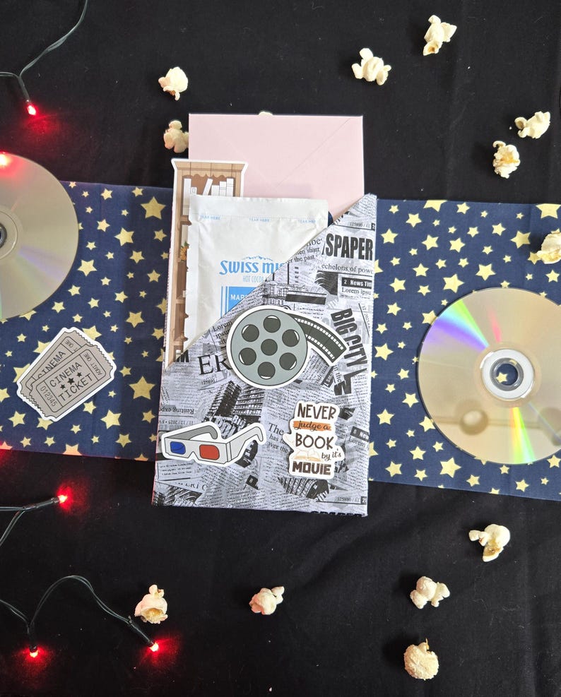 May include: A movie night gift set with a navy blue and gold star pattern, popcorn, and a CD. The set includes a pocket with newspaper print, a film reel sticker, and the text "NEVER judge a BOOK by its MOUIE".