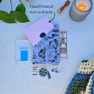 May include: A flat lay featuring a pink envelope sealed with a navy wax seal, a newspaper-print journal, and a packet of Swiss Miss marshmallows. Decorative elements include a lit candle, a wax stamp, and movie-themed stickers.