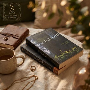 May include: Two books titled "the named" and "the dark" are stacked on a beige surface. The top book has a forest scene on the cover. A brown leather journal, a gold ring, a gold necklace, and a mug of coffee are also visible.