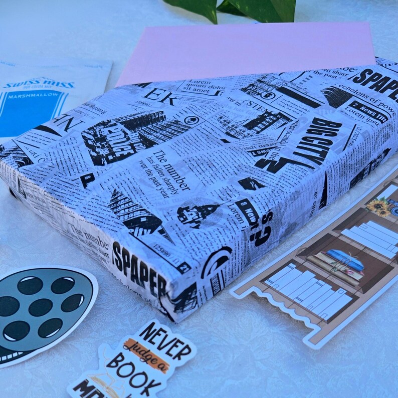 May include: A rectangular package wrapped in newspaper-style paper with black text and graphics. The package is surrounded by stickers, including one that says "Never judge a book". A film reel sticker and a Swiss Miss Marshmallow packet are also visible.