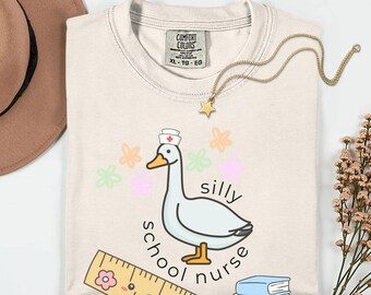 May include: Cream-colored t-shirt featuring a cartoon goose wearing a nurse's hat, with the words "silly school nurse." The design includes colorful flowers, a ruler, and a book. A gold necklace with a star pendant is also visible.