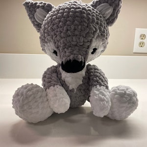 以下が含まれることがあります： グレーと白のハンドメイドかぎ針編みのオオカミのぬいぐるみ。オオカミはふわふわした質感で、耳、足、胸に白いアクセントがあります。目と鼻は黒です。ぬいぐるみは直立しています。