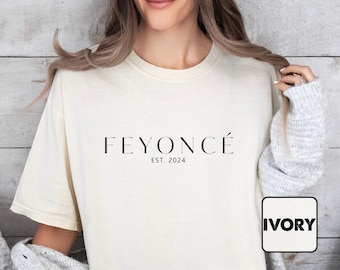 Può includere: Una donna indossa una maglietta color avorio chiaro con la scritta "FEYONCÉ EST. 2024" stampata sul davanti. La maglietta è di taglio ampio e ha un collo rotondo.