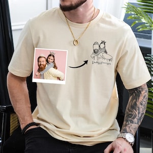 Może przedstawiać: Kremowy t-shirt z małym zdjęciem ojca i córki, rysunkiem linii tego samego obrazu i napisem "Daddy's Princesses". Koszulka ma krótkie rękawy i okrągły dekolt.
