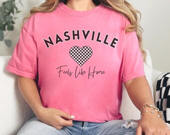 May include: Pink t-shirt with the text "Nashville Feels Like Home" in black. A black and white checkered heart is above the text.