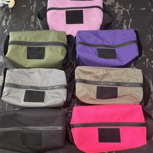 Puede incluir: Seis bolsas con cremallera de diferentes colores, incluyendo rosa, verde, morado, gris, negro y rosa brillante. Cada bolsa tiene un parche de velcro negro en la parte delantera.