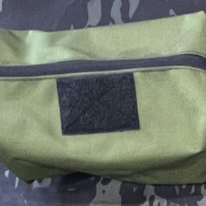 Puede incluir: Una bolsa de lona verde con cremallera, una correa negra y un parche de velcro negro.