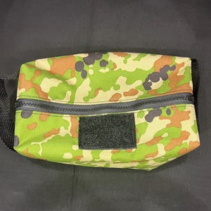 May include: A rectangular toiletry bag featuring a camouflage pattern in shades of green, tan, and dark brown. The bag includes a black zipper closure across the top, a black fabric handle on the side, and a rectangular black hook-and-loop patch.