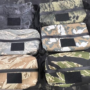 May include: Six camouflage-patterned pouches with black zippers and Velcro patches. The pouches feature various designs, including tiger stripe, digital, and woodland camouflage patterns. Each pouch has a black strap.