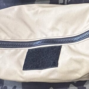 Puede incluir: Una bolsa de lona beige con cremallera, con una cremallera negra y un parche negro en la parte delantera.