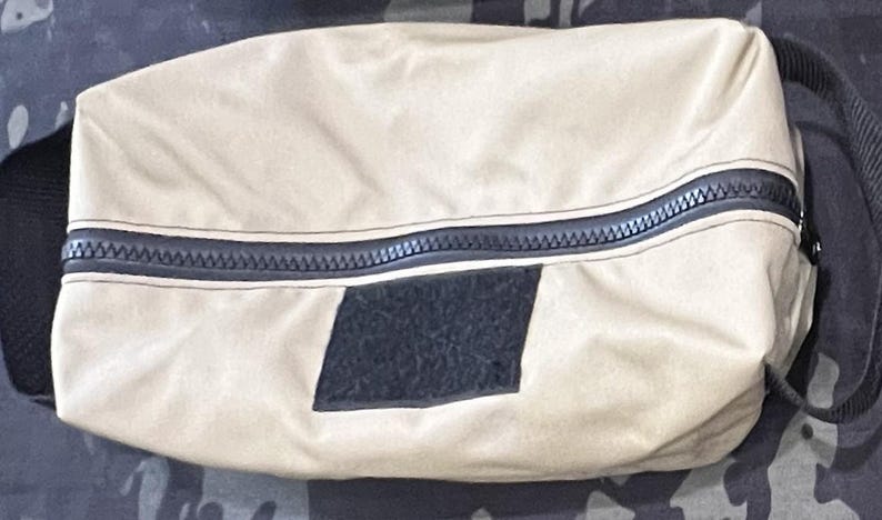 Puede incluir: Una bolsa de lona beige con cremallera, con una cremallera negra y un parche negro en la parte delantera.