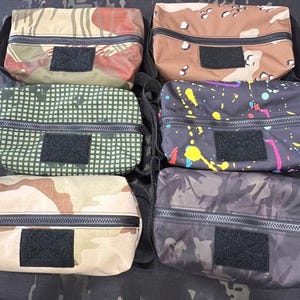 May include: Six different patterned zippered pouches with black zipper pulls and black velcro closures. The pouches are made of a variety of fabrics, including camouflage, paisley, and geometric patterns.