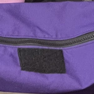 Puede incluir: Una bolsa de color morado con cremallera y un parche negro en la parte delantera. La bolsa est&aacute; hecha de un tejido suave y duradero y tiene un cierre de cremallera.