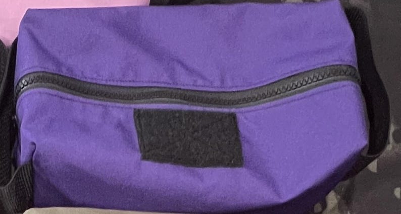 Puede incluir: Una bolsa de color morado con cremallera y un parche negro en la parte delantera. La bolsa est&aacute; hecha de un tejido suave y duradero y tiene un cierre de cremallera.