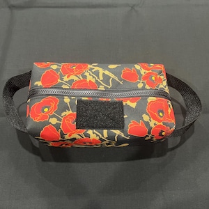 May include: A rectangular black pouch with a red poppy floral pattern and a black zipper. The pouch has a black loop handle and a black velcro patch. The background is a dark gray fabric.