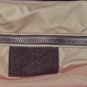 Puede incluir: Una bolsa beige con cremallera y un parche negro en la parte delantera. La bolsa tiene una cremallera negra y una correa negra.