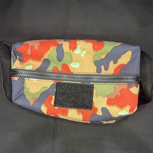 May include: A rectangular waist bag featuring a multi-colored camouflage pattern in shades of orange, olive green, tan, and dark blue. The bag includes a black horizontal zipper across the front and a rectangular black hook-and-loop patch.
