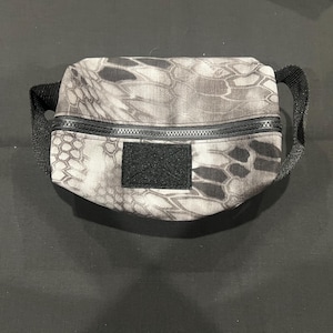 May include: A small, rectangular pouch with a black and gray camouflage pattern. The pouch has a black zipper closure and a black fabric handle. A black square patch is sewn on the front.