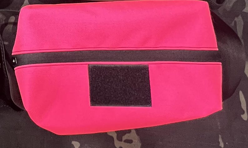 Puede incluir: Una bolsa de color rosa brillante con una cremallera negra y un parche de velcro negro. La bolsa est&aacute; hecha de un material suave y duradero y tiene una correa para llevar.