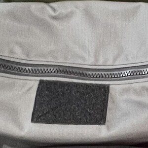 Puede incluir: Una bolsa gris con cremallera y un parche de velcro negro. La bolsa est&aacute; hecha de un material resistente al agua.