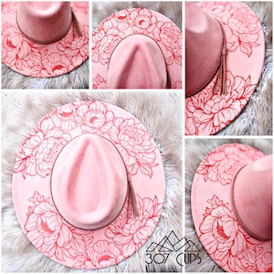 May include: A pink fedora-style hat with a wide brim, adorned with a red floral design. The hat features a matching pink crown, a brown hatband, and a tassel. The hat is displayed on a fluffy, white surface.