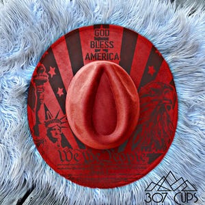 May include: A red fedora hat with a patriotic design. The hat features the text "God Bless America" and "We the People", along with images of the Statue of Liberty and an eagle. The hat is resting on a fluffy, light blue surface.