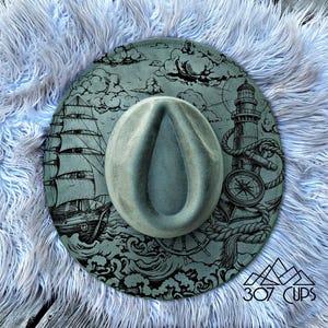 May include: A fedora hat with a nautical design. The hat features a ship, lighthouse, compass, and rope details in black on a teal background. The hat is displayed on a fluffy, light blue surface.