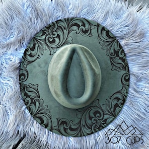 以下が含まれることがあります： つばに装飾的な黒いフローラルデザインが施された、明るいティール色のカウボーイハット。帽子はふわふわのライトブルーのフェイクファーの表面に置かれています。「307 CUPS」の文字が右下に表示されています。