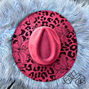 May include: A pink fedora-style hat with a wide brim featuring a black leopard print and floral design. The hat is set on a light blue, fluffy surface. The text "307 CUPS" is visible on the brim.