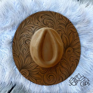 May include: A tan fedora-style hat with a brown, ornate floral design on the brim. The hat is set against a backdrop of white faux fur and a wooden surface. The text "307 CUPS" is visible in the lower right corner.