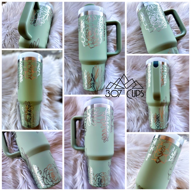 May include: A green travel mug with a silver lid and handle. The mug has a white floral design with a succulent theme. The mug is sitting on a white fluffy surface.